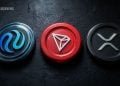 Three cryptocurrency coins laid in a row on a dark textured surface: blue logo on the left, a red triangular logo in the center, and a silver logo on the right (watermark says BLOCKNEWS).