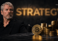 Businessman in a black suit sits beside Bitcoin and tall stacks of gold coins with a blurred 'STRATEGY' sign in the background.