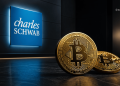 Gold Bitcoin coins on a dark floor with a blue Charles Schwab sign in the background.
