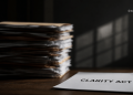 A tall stack of papers on a desk with a single sheet in the foreground reading 'CLARITY ACT' against a dark background.