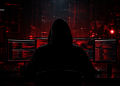 Hooded figure hunched over a desk with three monitors displaying red code in a dark red-lit room.