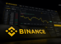 Binance logo in yellow on a dark surface with a blurred cryptocurrency price chart in the background