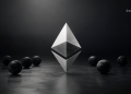 Metallic Ethereum logo standing upright with mirrored lower half, on a dark reflective surface with scattered black spheres.