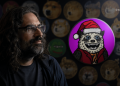 Man with long hair and glasses looks to the left beside a bright pink circular artwork of a cartoon wolf in a Santa hat. Background shows round decorative pieces and a BLOCKNEWS logo.