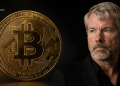 Large gold Bitcoin coin beside a serious man in a black suit against a dark background, suggesting crypto news or finance.