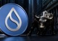 Large blue cryptocurrency coin with a white flame-like logo in front of a bronze Charging Bull statue on a city street.