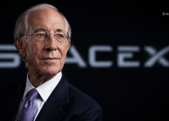 Portrait of an older man in a dark pinstripe suit and purple tie, looking to the left, with a blurred logo in the background.