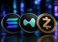 Three cryptocurrency coins—Dash (blue logo), Monero (teal logo), and Zcash (gold logo)—resting on a dark, reflective surface with neon-blue background lights.
