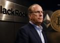 Businessman in a suit stands beside a BlackRock sign with a Bitcoin graphic in the background.
