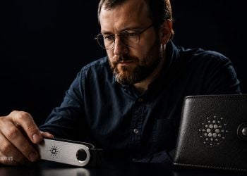 Man with glasses and beard uses a metal cryptocurrency hardware wallet on a dark table, with a black leather wallet beside him.