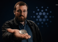 Bearded man with glasses in a leather jacket extends a silver cryptocurrency coin toward the camera against a dark background.