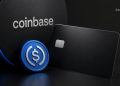 Blue circular coin with a dollar sign sits beside a dark laptop and a metal chip-enabled card, with the Coinbase logo in the background.