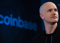 Close-up of a bald man in a black jacket looking slightly upward, standing on a dark blue stage backdrop with a blurred blue sign in the background (BlockNews watermark).