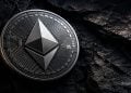 Close-up of a metallic Ethereum coin resting on a dark textured rock surface, showing circuit-like engravings and the Ethereum logo at the center.