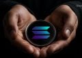 Person holds a glossy round crypto coin with a blue‑purple gradient geometric logo in cupped hands.