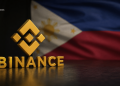 Gold Binance logo and letters on a dark surface with a blurred Philippine flag in the background