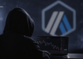 Person in a hoodie sits at a laptop displaying a stock/crypto chart in a dark room with a blue geometric logo on the wall far behind them