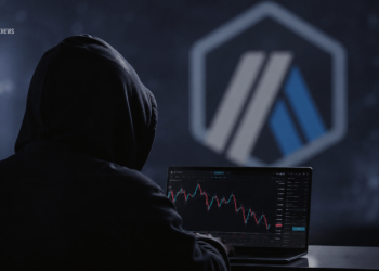 Person in a hoodie sits at a laptop displaying a stock/crypto chart in a dark room with a blue geometric logo on the wall far behind them