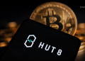 Close-up of a Bitcoin coin behind a tilted smartphone displaying the Hut 8 logo on a dark, blurred background.