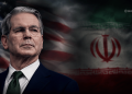 Portrait of a serious-looking man in a dark suit and green tie, with the American flag and the Iranian flag in a smoky backdrop behind him.